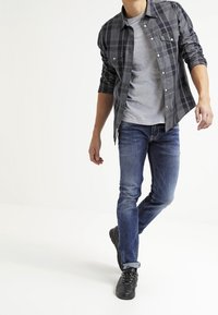 Camisa a cuadros gris y negra sobre una camiseta gris, vaqueros azules y zapatillas negras. La camisa tiene detalles de botones y mangas remangadas.