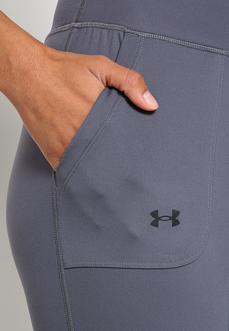 Un pantalon de sport gris avec une poche latérale, des coutures surpiquées et le logo Under Armour en noir près de la poche. Le matériau semble lisse.