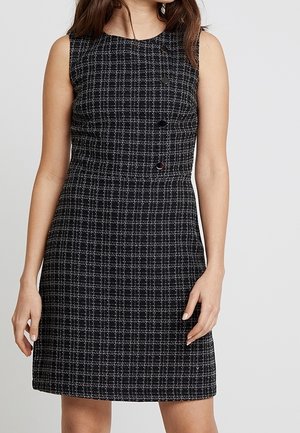 Černobílé kostkované šaty bez rukávů s asymetrickými knoflíky na horní přední straně, které nosí stojící žena na jednobarevném pozadí.