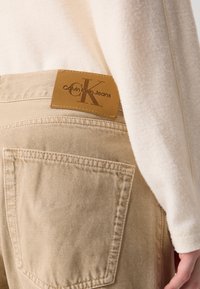 Jeans di denim cotone color sabbia presentano una tasca posteriore rettangolare e un'etichetta in pelle con il logo "Calvin Klein Jeans". Il tessuto ha una superficie liscia.