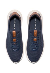 Námořnické modré tenisky s texturovaným pleteným svrškem a hnědými koženými akcenty. Mají kulatou špičku a plochou bílou podrážku. Šňůrky jsou umístěny uprostřed.