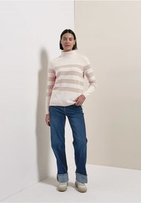 Pull à col roulé rayé rose clair et blanc en matière douce, associé à un jean bleu et des baskets beige. Le mannequin pose devant un fond neutre.