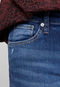 Vaqueros de mezclilla azul oscuro con un primer plano del bolsillo delantero y los detalles de la cinturilla. Presenta costuras visibles y una zona desgastada.