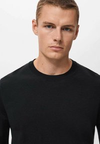 Črna pletena jopica s krožnim izrezom, ki ima gladko teksturo in prilegajoč dizajn, minimalni šivi ob izrezu in robu.