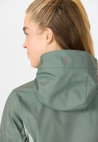 Grön huva jacka med ett slätt, vattenavvisande tyg. Har en hög krage och subtil logotypdetalj nära axelsömmen.