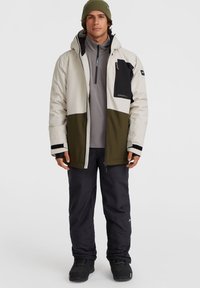 Veste de neige pour homme en crème, vert olive et noir, dotée d'une poche zippée, de poignets ajustables, et associée à un pantalon sombre et des bottes.