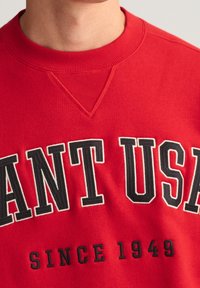 Sudadera roja con cuello redondo acanalado, "ANT USA" en letras negritas negras, con "SINCE 1949" debajo en texto negro más pequeño. Textura suave.