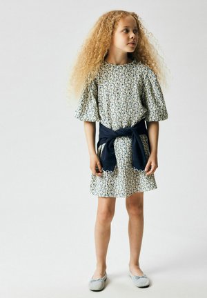 Nuori tyttö, jolla on pitkät kiharat hiukset ja kukkamekko, laivastonsininen neule solmittuna vyötärölle ja harmaat balettikengät, seisoo yksinkertaisen taustan edessä.