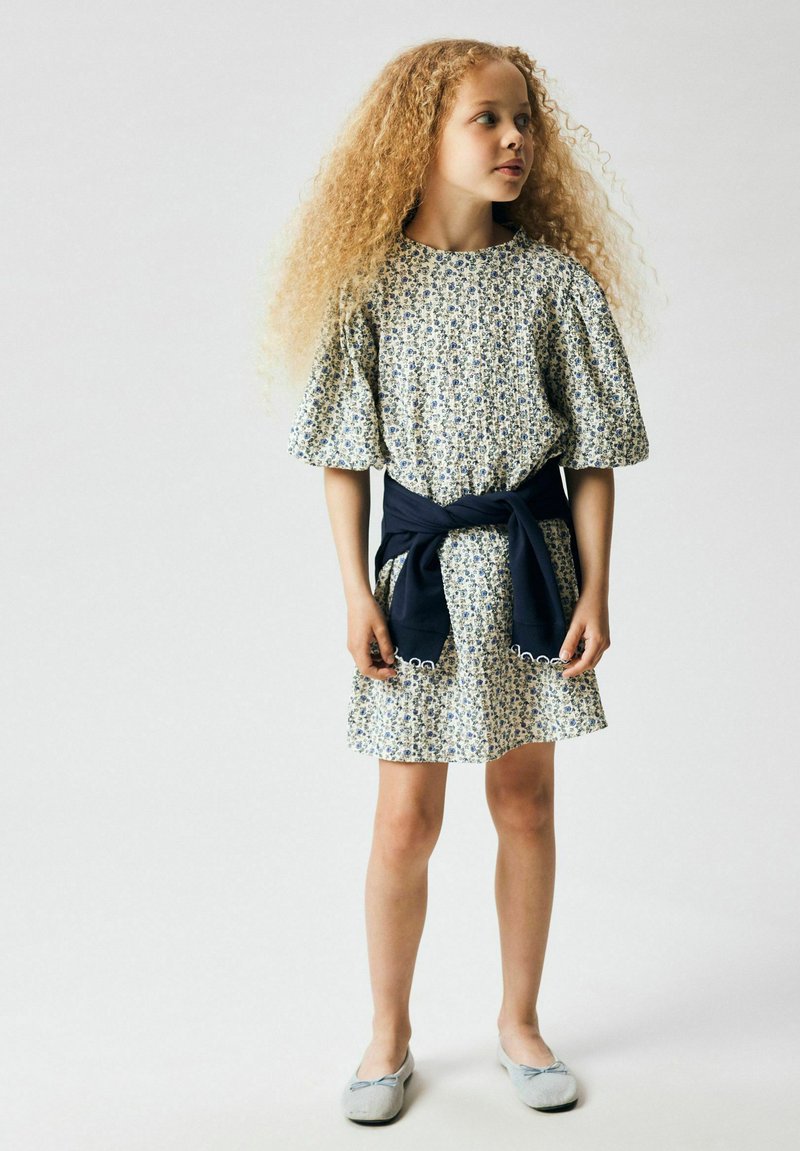 Jeune fille aux longs cheveux bouclés portant une robe à fleurs, un pull marine noué à la taille, et des ballerines grises, debout devant un fond uni.