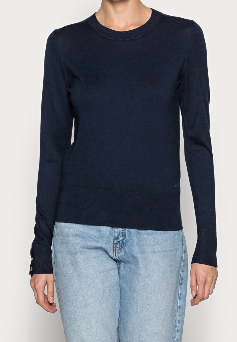 Femme portant un pull ajusté bleu marine à manches longues avec des détails de boutons aux poignets et un jean en denim bleu clair, debout devant un fond uni.