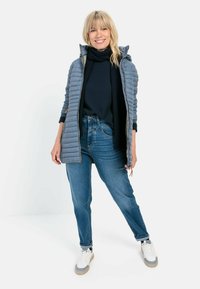 Grijze pufferjack met rits en capuchon, gedragen over een zwarte trui en blauwe spijkerbroek. Geaccentueerd met een zwarte sjaal en witte sneakers.