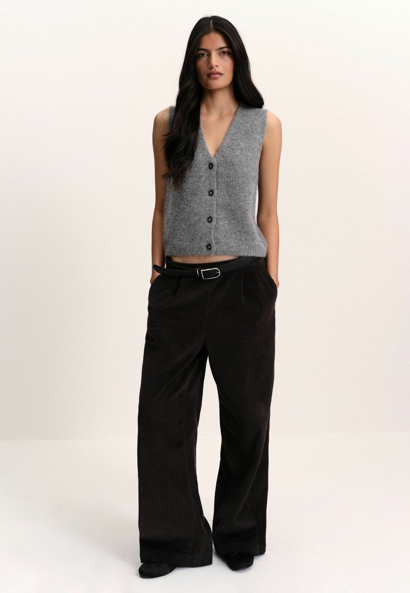 Woman with long dark hair wearing a gray sleeveless buttoned vest, black wide-leg pants with belt, standing with hands in pockets.