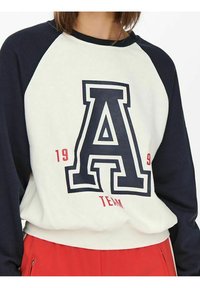 Sudadera de manga larga en blanco y azul marino que presenta un gran gráfico en forma de "A" con detalles en rojo y una textura suave. Diseño casual y ajustado.