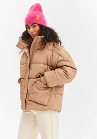 Hnedá prešívaná vatovaná bunda s vysokým golierom a bočnými vreckami, v páre s jasnoružovou pletenou čiapkou, na prednej strane s visačkou.