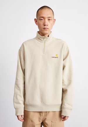 Beige fleece pullover with a half-zip collar, featuring a small embroidered logo in red and yellow. It has ribbed cuffs and hem.