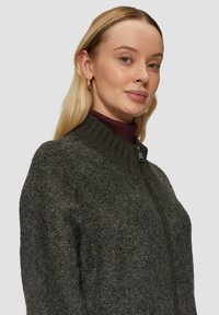 Donkergrijze zip-up jas met hoge kraag, gestructureerde stof en minimale hardware. Gelaagd over een bordeauxrode coltrui.