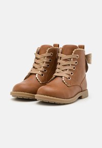 Bottes chevilles en cuir marron avec des lacets dorés, des bords festonnés et un accent de nœud scintillant à l'arrière. Semelle en caoutchouc solide pour plus de durabilité.