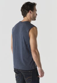 Hombre con camisa sin mangas azul marino descolorida y vaqueros oscuros, de pie de perfil frente a un fondo claro liso.