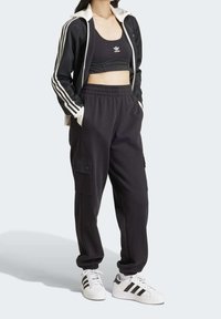 Černá sportovní souprava zahrnující bundu na zip s bílými pruhy, přiléhavý krátký top a cargo kalhoty s bočními kapsami, doplněná bílými teniskami.