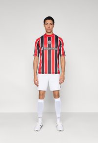 Punaste ja mustade triipudega jalgpallimärk, ümar kaelus, logoga ja sponsori tekstiga, valgete lühikeste pükste ja sokkidega.