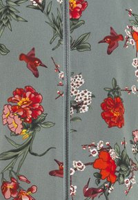 Szary materiał z srebrnym zamkiem błyskawicznym, ozdobiony czerwonymi i pomarańczowymi kwiatami, białymi kwiatami, zielonymi liśćmi oraz małymi czerwonymi koliberami.