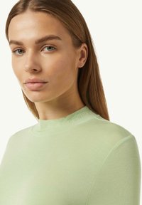 Lichtgroene langemouwen top met een hoge halslijn, gemaakt van gladde stof. Heeft reliëftekst op de kraag die "CASUAL IDENTITY" zegt.