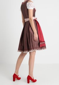 Tenue traditionnelle avec un corsage ajusté et une jupe évasée, composée d'un tissu marron à motif, d'un accent rouge et d'un haut blanc en dentelle. Talons rouges.