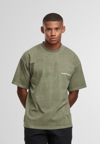 Mladý muž s krátkými vlasy, oblečený v olivově zeleném oversized tričku s nápisem "Sunday Running Club" na hrudi, stojící proti jednobarevnému světlému pozadí.