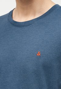 Κοντινό πλάνο ενός ατόμου που φοράει μπλε μπλούζα με στρογγυλή λαιμόκοψη και ένα μικρό πορτοκαλί λογότυπο αμπερσάντ κεντημένο στο αριστερό μέρος του στήθους.