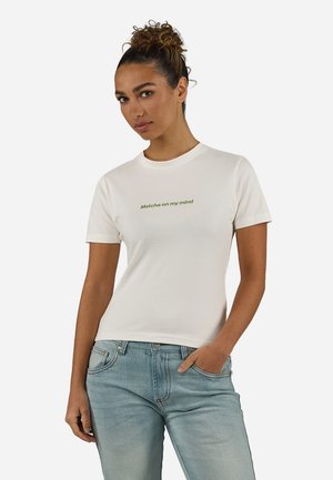 Γυναίκα με σγουρά μαλλιά δεμένα, φοράει λευκό μπλουζάκι με πράσινο κείμενο "Matcha on my mind" και ανοιχτόχρωμο τζιν, στέκεται με το ένα χέρι στην τσέπη.