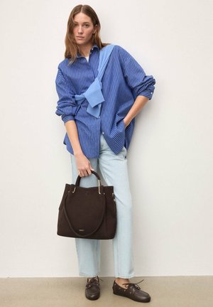 Ženska, oblečena v modro srajco z črtami in svetle kavbojke, drži veliko temno rjavo ročno torbo, čez ramena pa ima premeten svetlo moder pulover.