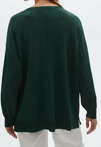 Maglione oversize verde scuro con scollatura a barca, maniche lunghe e spacchi laterali all'orlo. Tessuto liscio, vestibilità comoda, dettagli minimi.