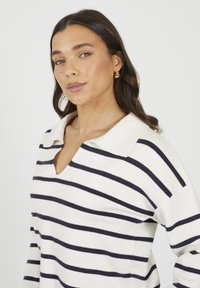 Kobieta z długimi ciemnymi włosami, nosząca złote kolczyki w kształcie kół oraz białą sweter z kołnierzykiem w czarne poziome paski, pozująca na białym tle.