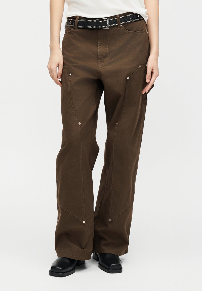 Άτομο που φοράει καφέ παντελόνι φαρδιάς γραμμής με ασημένιες τρουκς και μαύρη ζώνη με τρουκς σε σχήμα αστεριού, σετάρισμα με μαύρα παπούτσια και λευκό τοπ.