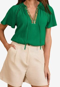Top verde a maniche corte con scollatura a V, caratterizzato da dettagli in paillettes dorate e accenti texturizzati. Abbinato a pantaloni corti beige su misura.