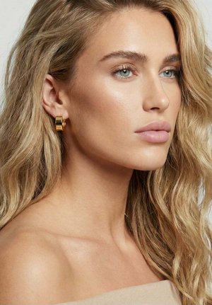 Lyshåret kvinde med bølget hår, iført guldringe, kigger mod højre med neutralt udtryk og bare skuldre synlige.