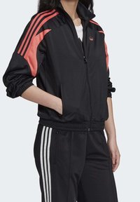 Svart zip-up jacka med rosa detaljer, tillverkad av slätt tyg. Har tre ränder på ärmarna och elastiska muddar, med sidofickor.