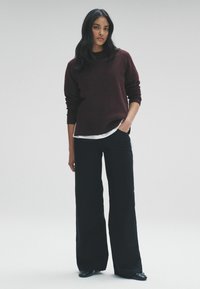 Burgundský pletený svetr přes bílou košili, doplněný o černé kalhoty s širokými nohavicemi a černé boty. Model stojí s rukama v kapsách.