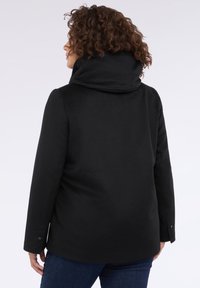 Cămașă neagră cu un guler înalt, fabricată dintr-un material moale. Are un croi drept și mâneci lungi cu accente de nasturi la manșete.