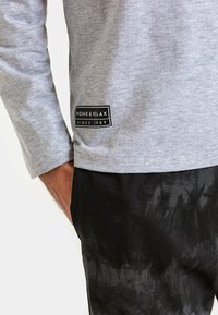 Szara koszula z długim rękawem z czarną prostokątną naszywką z napisem "DOM I RELAKS OD 1989 R." Noszona z czarnymi, strukturalnymi spodniami.