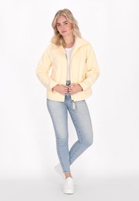 Jauna moteris pasitiki savimi stovėdama šviesiai geltonoje striukėje, baltoje marškinėliuose ir šviesiai mėlynuose džinsuose, prieš paprastą baltą fono.