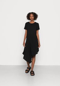 Vestido preto assimétrico com mangas curtas, tecido drapeado e uma textura suave. Combinado com sandálias chunky pretas, em pé sobre um fundo neutro.