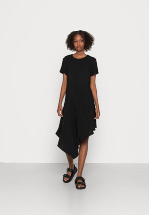 Vestido preto assimétrico com mangas curtas, tecido drapeado e uma textura suave. Combinado com sandálias chunky pretas, em pé sobre um fundo neutro.