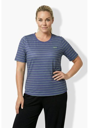 Frau mit blondem Haar, die ein gestreiftes kurzärmeliges Hemd in Blau und Grün sowie eine schwarze Hose trägt und mit einer Hand an der Hüfte steht.