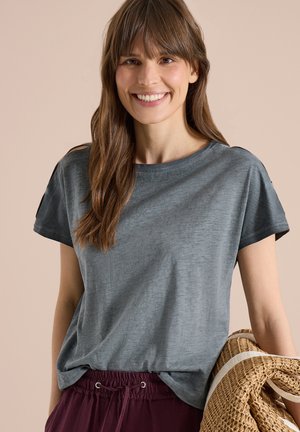 Lächelnde Frau mit langen braunen Haaren, die ein graues T-Shirt und bordeauxrote Hosen trägt und eine zusammengerollte gewebte Matte mit einem weißen Streifen hält.