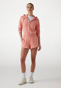 Haut à capuche rose zippé avec cordons, shorts assortis, le tout en tissu doux. Graphisme blanc sur le haut à capuche. Baskets blanches et chaussettes de cheville.