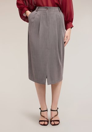 Donna che indossa una gonna grigia a vita alta, lunga fino al ginocchio con pieghe frontali, abbinata a una blusa rossa trasparente e tacchi neri con cinturini.