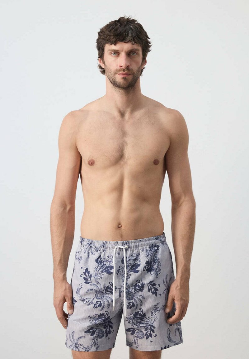 Homme torse nu aux cheveux courts et bouclés portant un short de bain clair à motif floral avec un cordon blanc, debout devant un fond uni.
