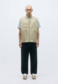 Μπεζ αμάνικο γιλέκο με φερμουάρ μπροστά, διακοσμημένο με πράσινη λεπτομέρεια, φορεμένο πάνω από ένα ανοιχτό μπλε πουκάμισο και συνδυασμένο με σκούρους παντελόνες και αθλητικά παπούτσια.