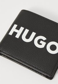 Cartera de cuero negra con "HUGO" en letras blancas en negrita, textura granulada y bordes cosidos. Diseño compacto y de doble pliegue.